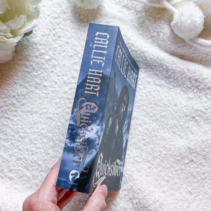 A hand holds a book, featuring a dark blue cover with a hooded figure, set against a soft white textured blanket background with white flowers.