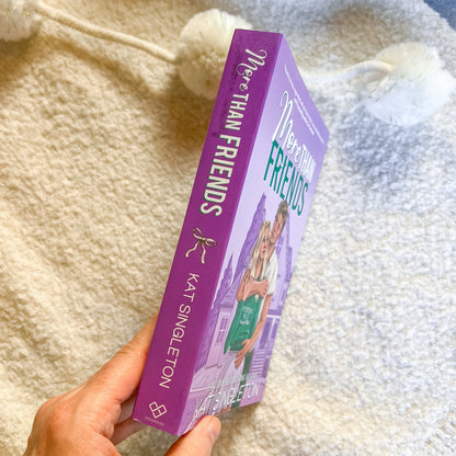 Purple book titled 'More Than Friends' by Kat Singleton held against a soft, textured background.