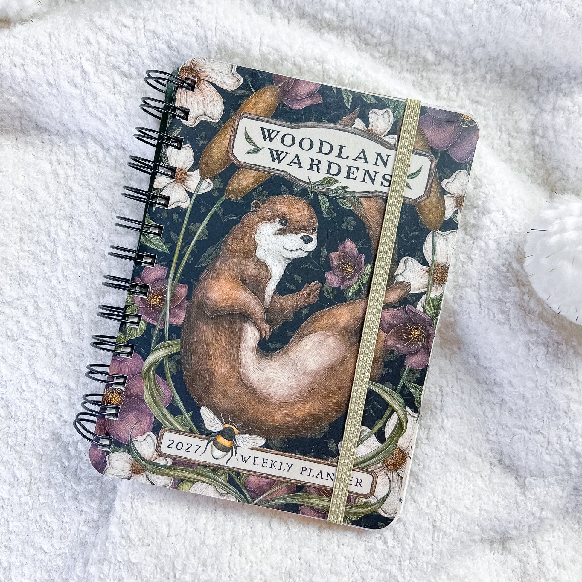 A spiral-bound 2027 weekly planner for 'Woodland Wardens' featuring an illustration of an otter on the cover, styled on a white textured blanket.