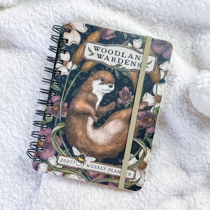A spiral-bound 2027 weekly planner for 'Woodland Wardens' featuring an illustration of an otter on the cover, styled on a white textured blanket.