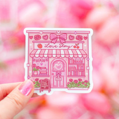 Pink 'Love Story' building sticker held by a hand with a blurred pink background