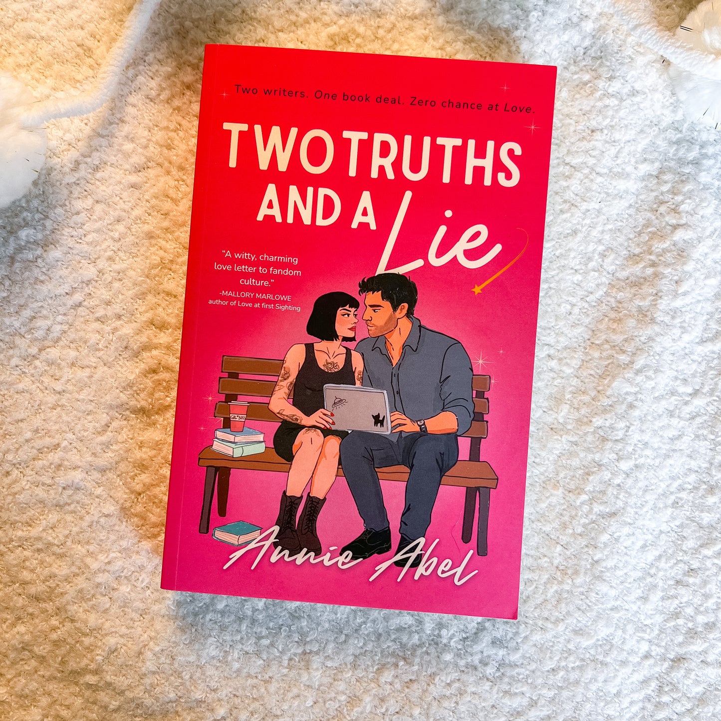A book titled 'Two Truths and a Lie' by Annie Abel, featuring a bright pink cover with an illustration of a couple sitting on a bench using a laptop, placed on a white textured blanket.