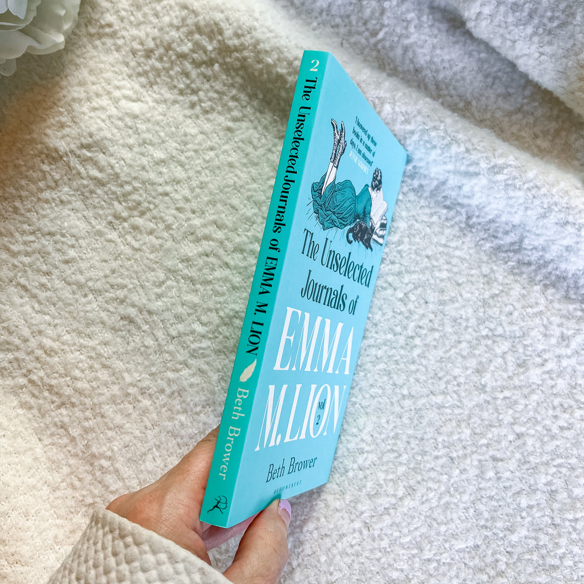 A hand holding a light blue book titled "The Unselected Journals of Emma W. Lion" by Beth Browner, photographed against a soft white textured blanket background.