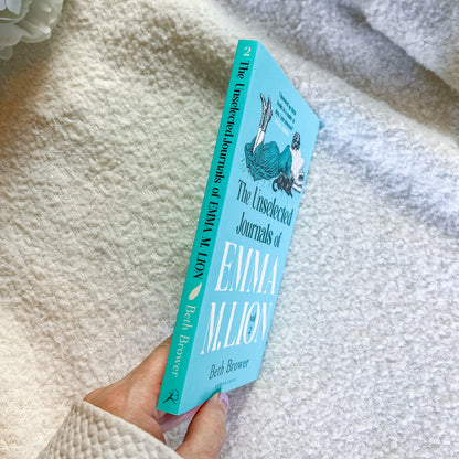 A hand holding a light blue book titled "The Unselected Journals of Emma W. Lion" by Beth Browner, photographed against a soft white textured blanket background.