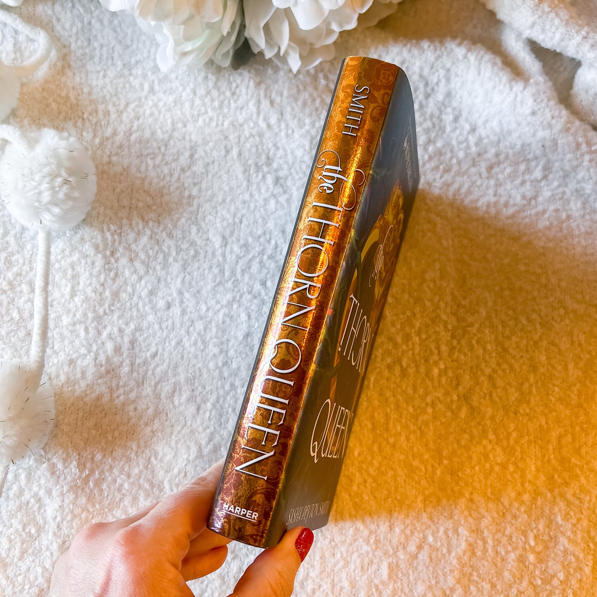 Book titled 'The Thorn Queen' held by a hand on a textured surface with flowers in the background