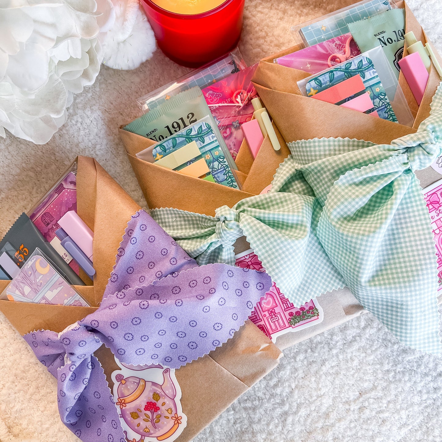 Three handmade gift wrapped blind date books made of brown kraft paper, tied with decorative fabric bows in purple, mint green, and light blue gingham. The pouches are filled with colorful stationery items and are styled on a soft cream-colored blanket next to a lit red candle and white flowers.