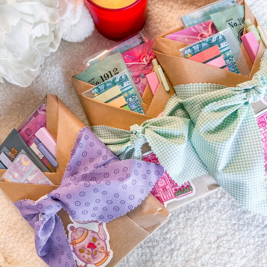 Three handmade gift wrapped blind date books made of brown kraft paper, tied with decorative fabric bows in purple, mint green, and light blue gingham. The pouches are filled with colorful stationery items and are styled on a soft cream-colored blanket next to a lit red candle and white flowers.