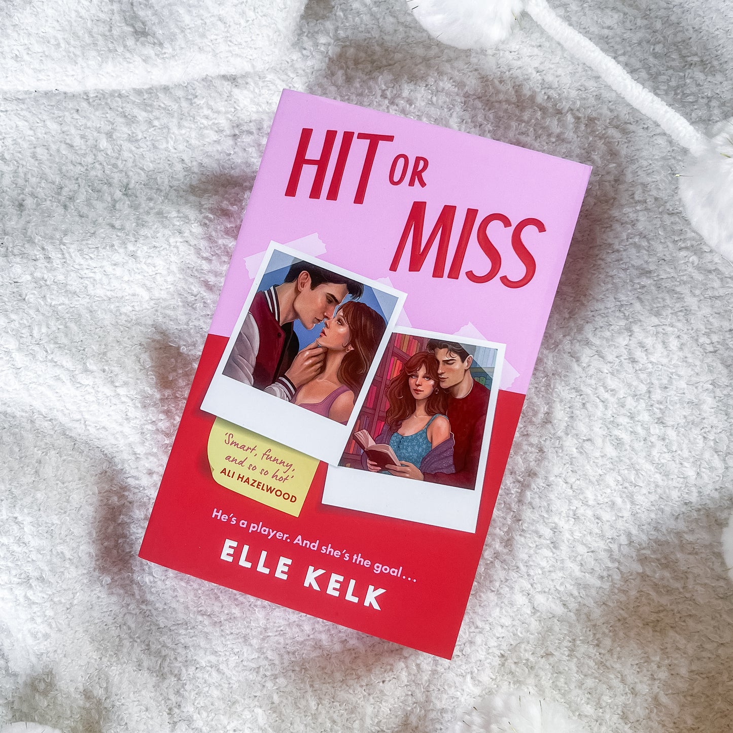 The book cover of 'Hit or Miss' by Elle Kelk is displayed diagonally on a soft, white textured blanket. The cover features a pink top half with bold red text, a red bottom half, and two polaroid-style photos showing a couple.