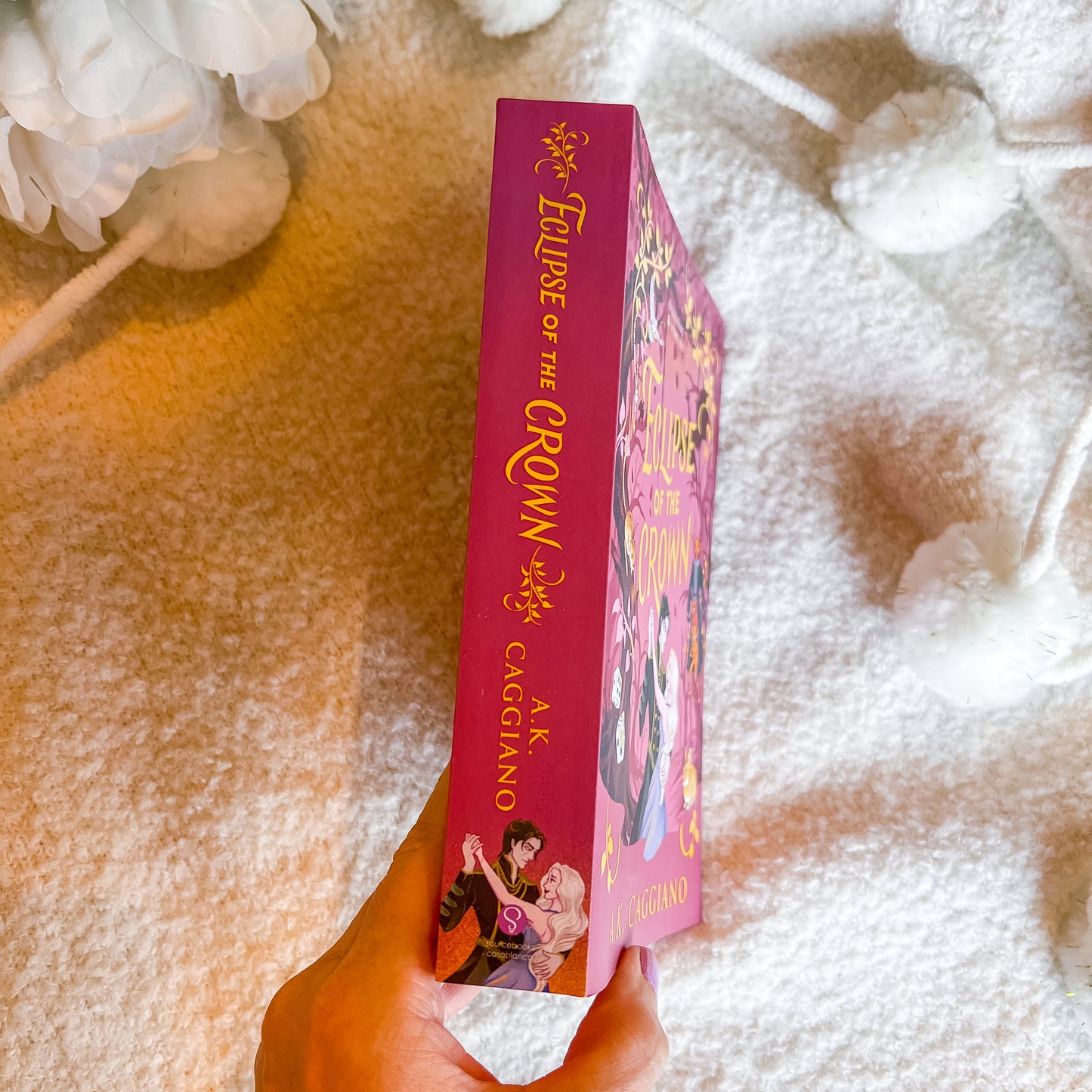 A hand holds a pink hardcover book against a soft white textured blanket background with decorative white flowers.