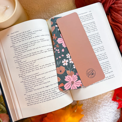 A rectangular rose gold bookmark with a floral pattern and a circular logo sits on the pages of an open book, styled with a candle, dried flowers, and autumn leaves.