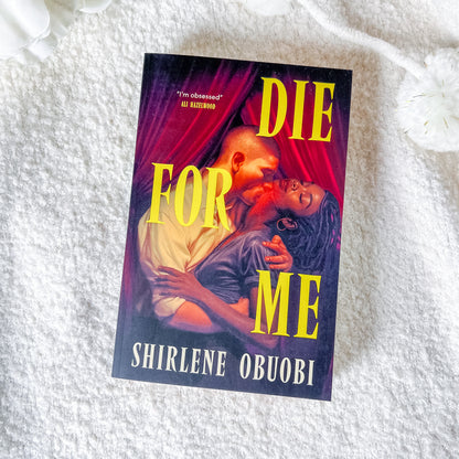A book titled 'Die For Me' by Shirlene Obuobi is displayed flat on a textured white blanket, with white flowers as decorative props in the corners. The cover art features a romantic embrace between a man and a woman under a deep red curtain, with bold yellow title text and white author text.