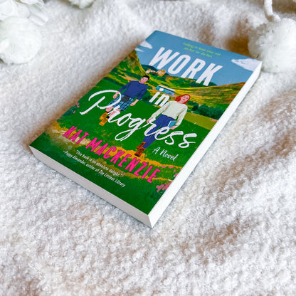 A paperback book titled "Work in Progress"  lies on a soft white textured blanket. The cover features a colorful illustration of two people walking through a green field under a blue sky.