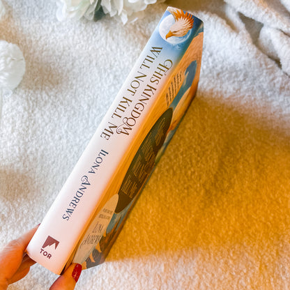 Book titled 'This Kingdom Will Not Kill Me” by Lona Andrews held by a hand on a textured surface.