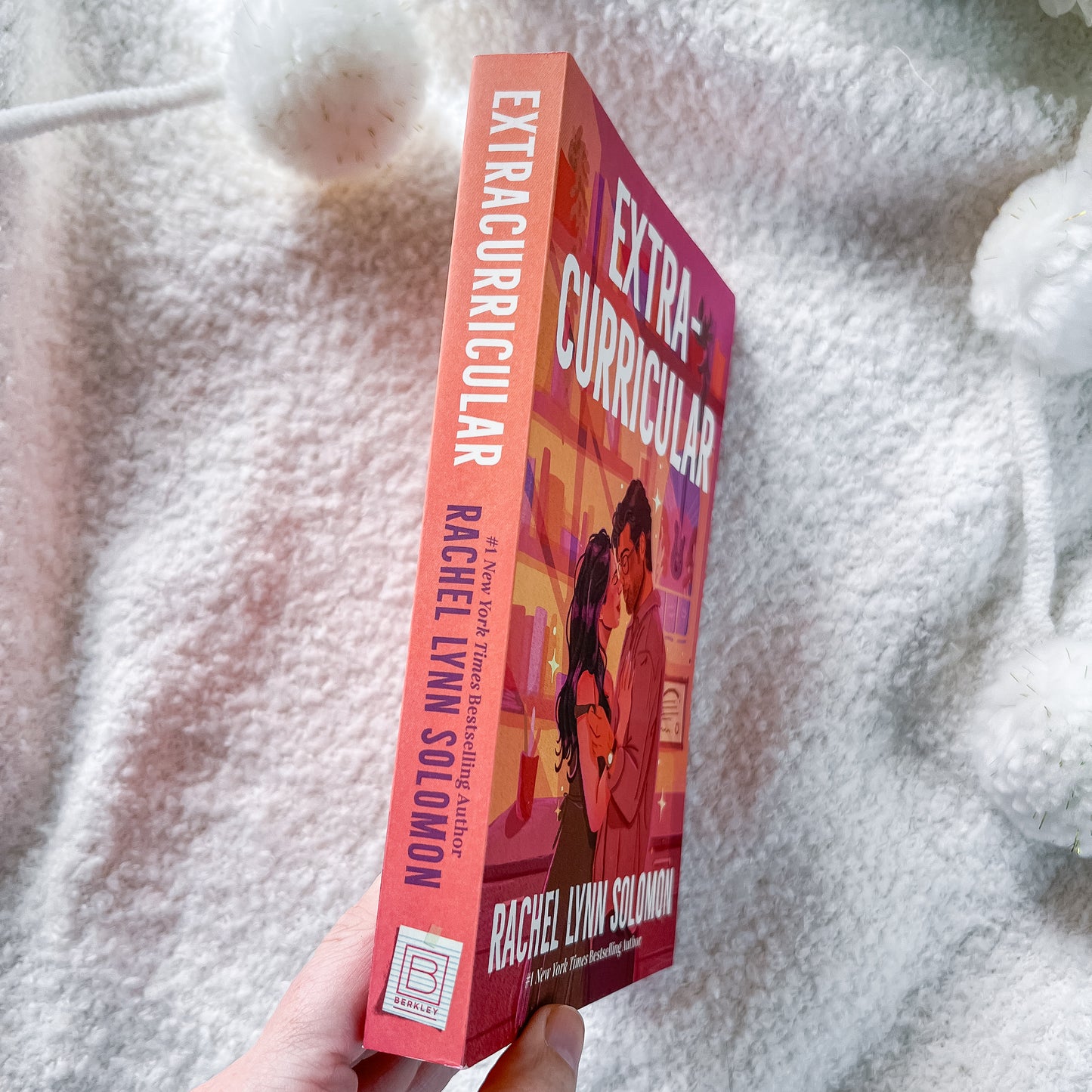 A hand holds a copy of the book 'Extra-Curricular' by Rachel Lynn Solomon, showing the front cover and spine, set against a soft, white fuzzy blanket background.