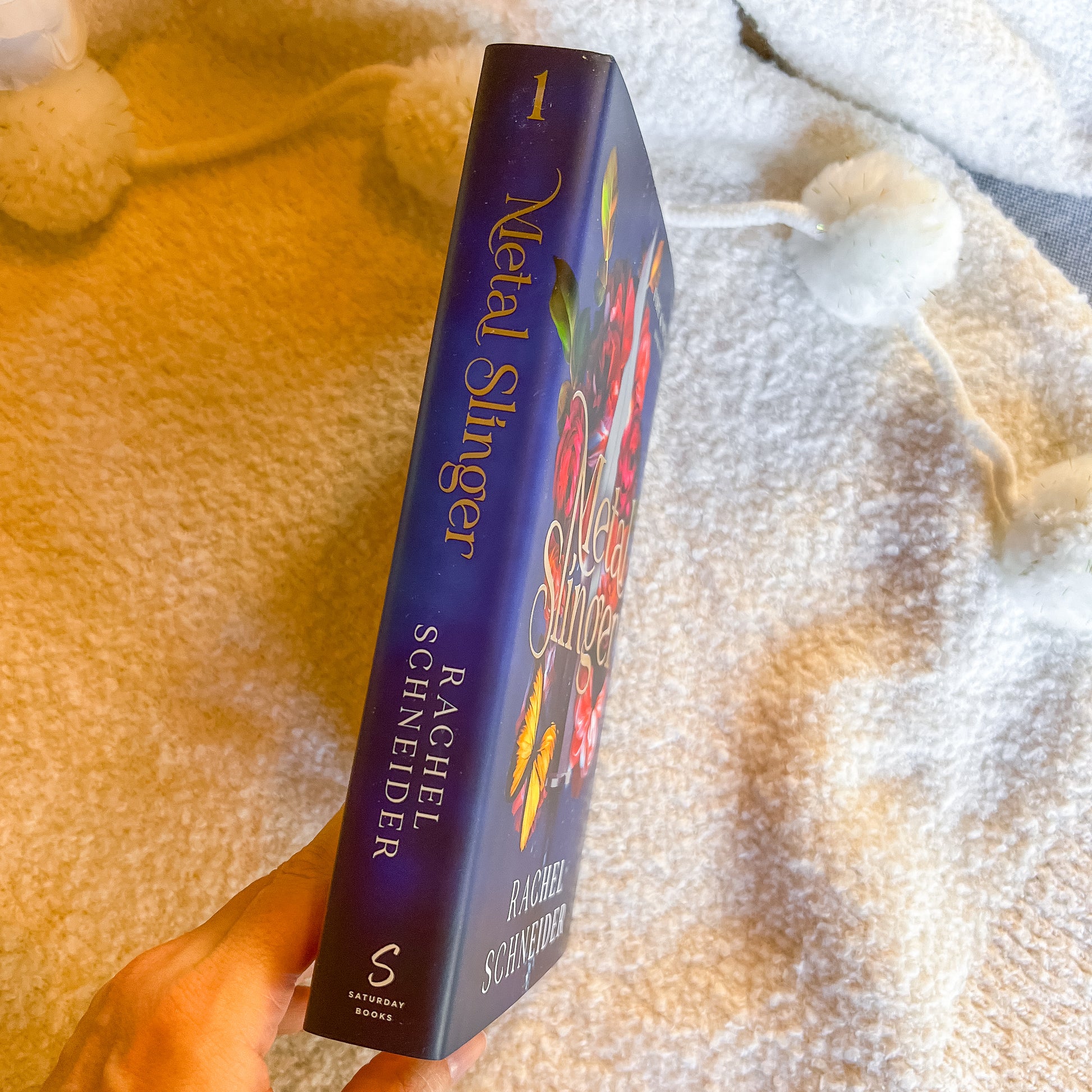 Book titled 'Metal Slinger' by Rachel Schneider held against a textured fabric background