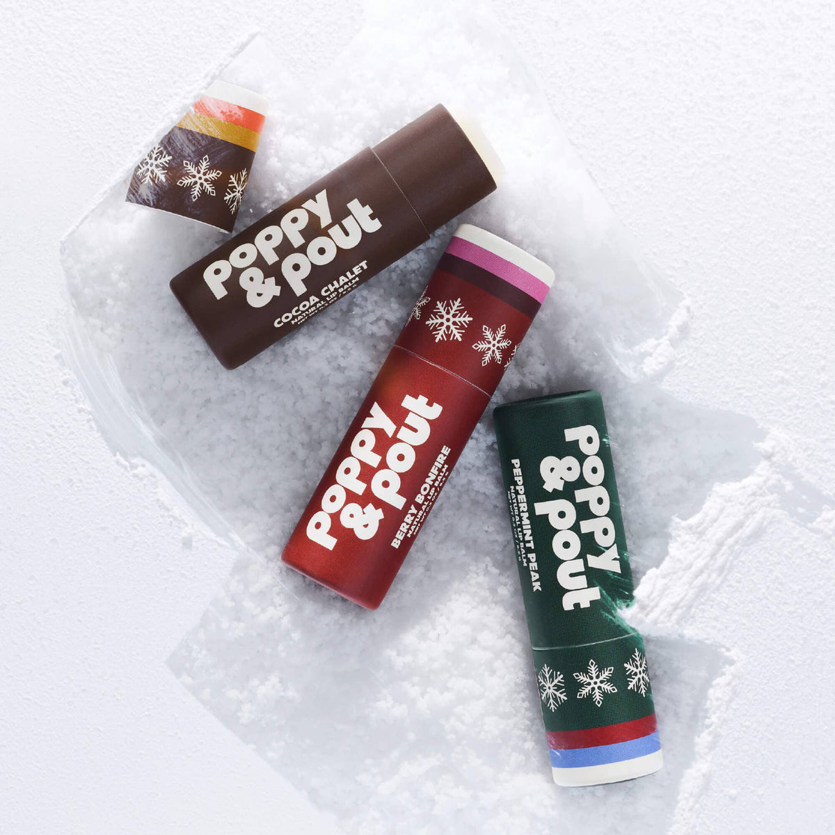 Three Poppy & Pout lip balm tubes arranged on white snow. The tubes are brown, red, and green with white text and snowflake designs, labeled Cocoa Chant, Red Powder, and Pepperoni Pie respectively.
