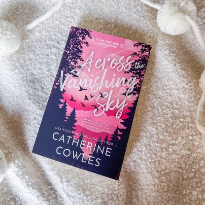 A paperback copy of the book 'Across the Vanishing Sky' by Catherine Cowles, featuring a pink and dark blue cover with silhouettes of trees and birds, placed on a soft white textured blanket.