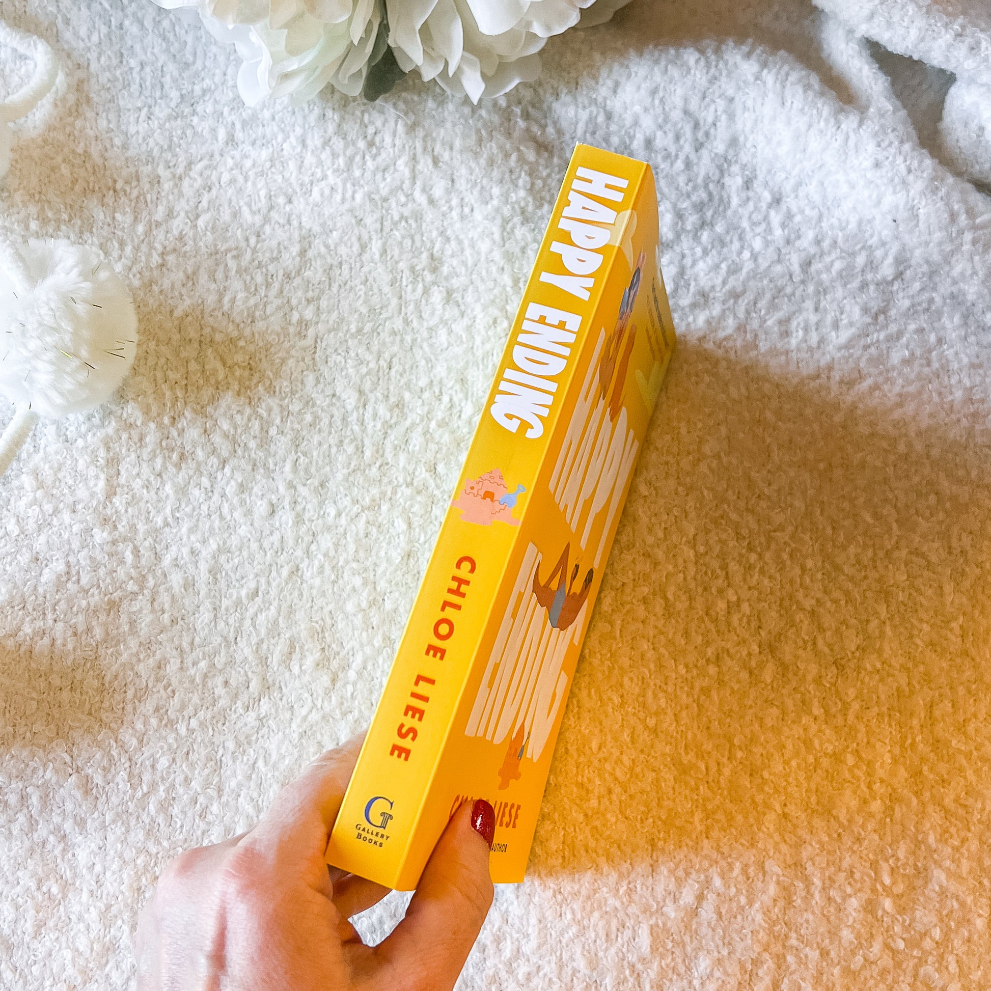Hand holding a yellow book titled 'Happy Ending' on a textured surface with flowers in the background