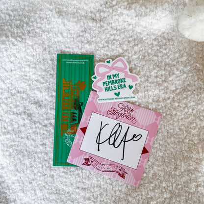 A flat-lay of a handmade paper ephemera set on a white textured rug. The set includes a green rectangular card with gold foil lettering, a pink bow-shaped sticker reading 'IN MY PEMBROKE HILLS ERA', and a pink striped card featuring the words 'Hot September' and a black ink signature.
