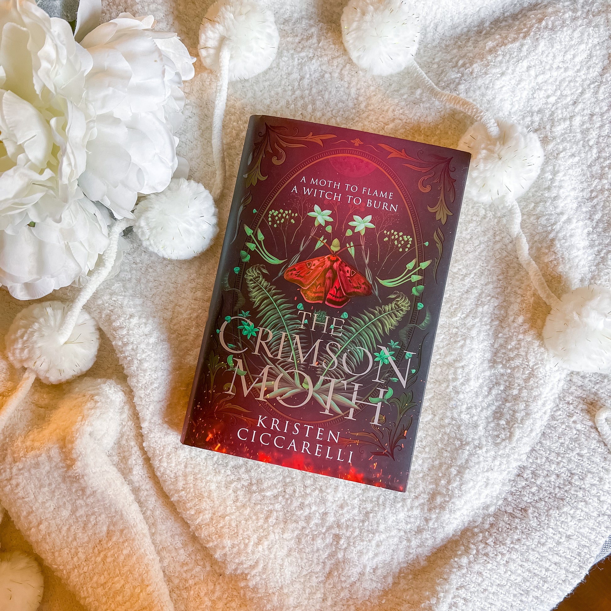Book titled 'The Crimson Moth' by Kristen Ciccarelli on a textured surface with white flowers.
