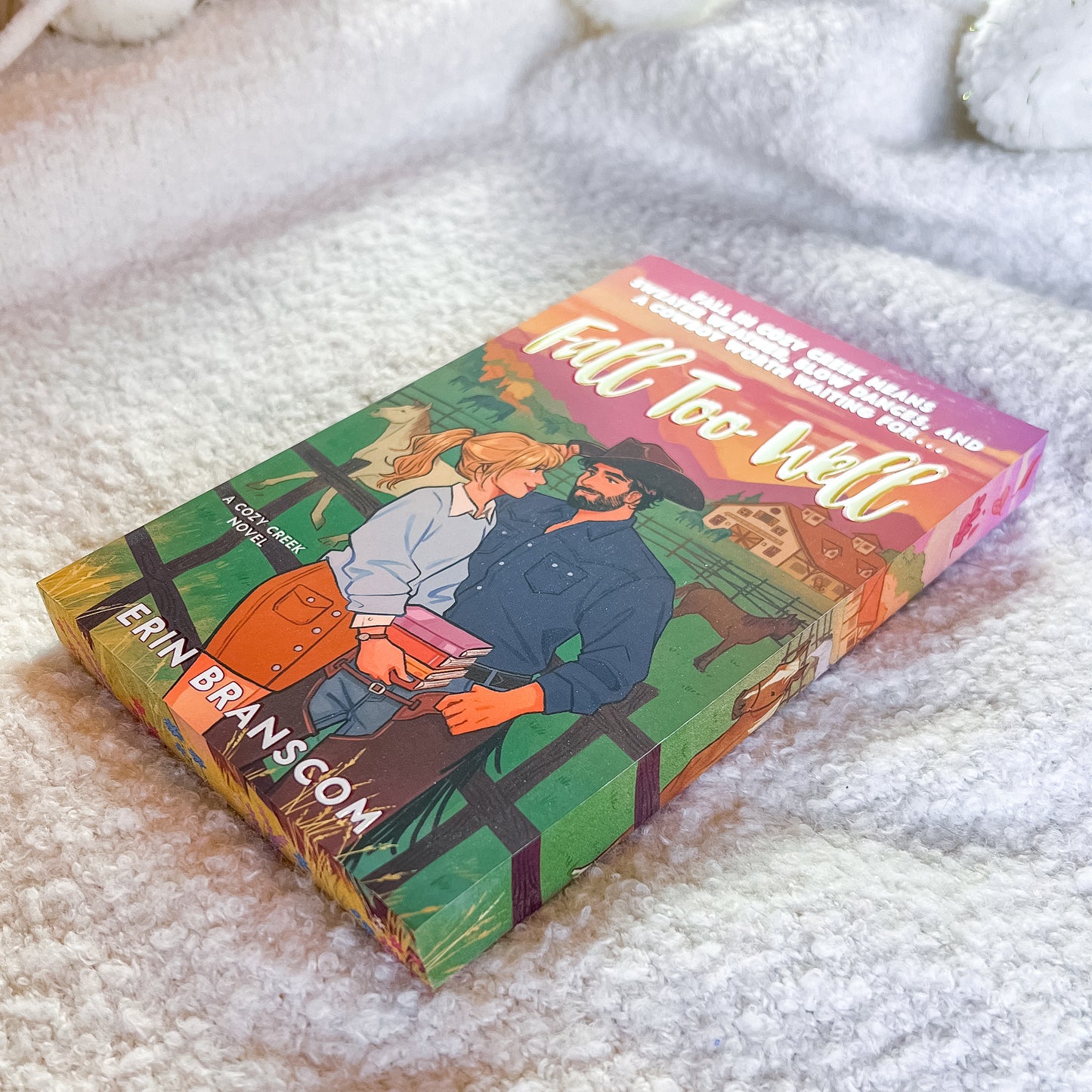 A book titled "Fall Too Well" by Erin Branscom lies on a soft white textured blanket. The cover features a colorful illustration of a couple embracing in front of a barn, with a pink and orange sunset sky.