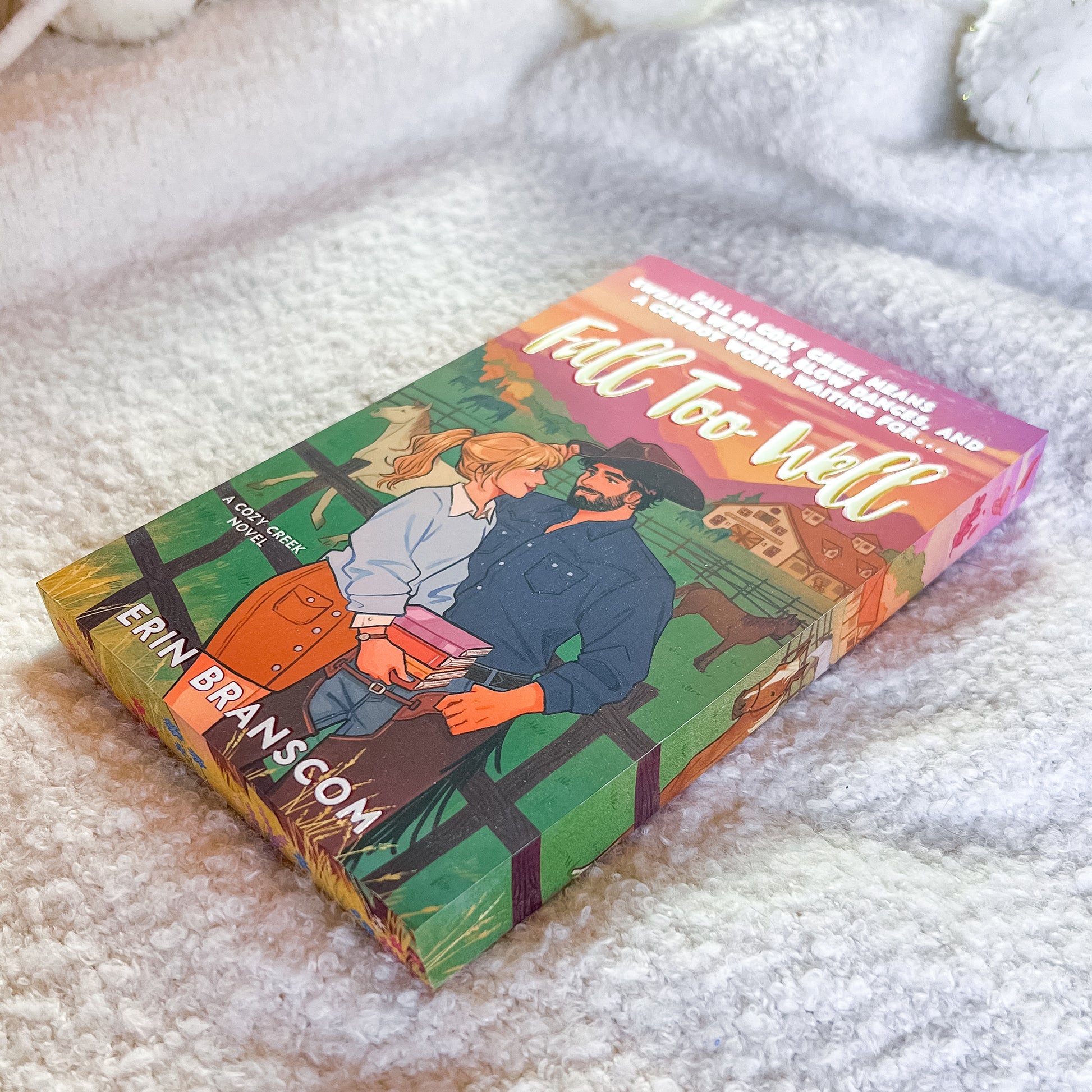 A book titled "Fall Too Well" by Erin Branscom lies on a soft white textured blanket. The cover features a colorful illustration of a couple embracing in front of a barn, with a pink and orange sunset sky.