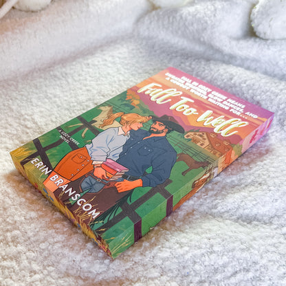 A book titled "Fall Too Well" by Erin Branscom lies on a soft white textured blanket. The cover features a colorful illustration of a couple embracing in front of a barn, with a pink and orange sunset sky.