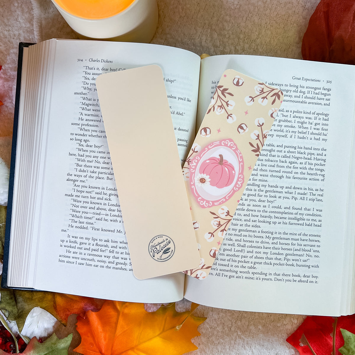 A decorative book bookmark with a pink pumpkin character and floral border, placed on an open book alongside a simple beige bookmark.