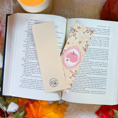 A decorative book bookmark with a pink pumpkin character and floral border, placed on an open book alongside a simple beige bookmark.