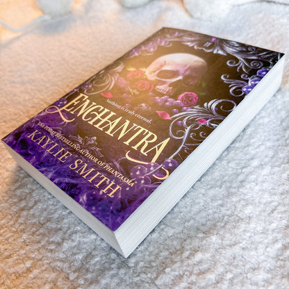 A book by Kaylie Smith, featuring a dark cover with a skull, roses, and purple ornate details, photographed on a soft white textured blanket.