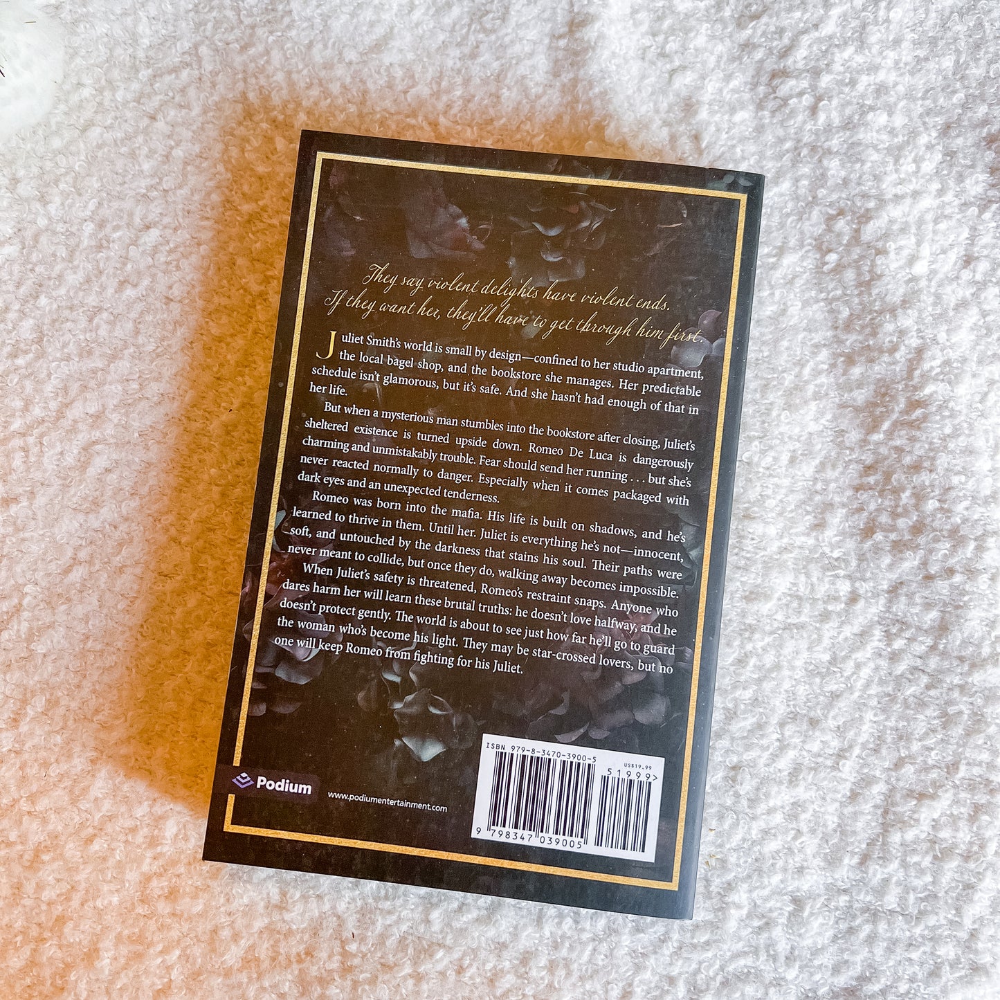 The back cover of a book by Podium, photographed flat on a textured beige carpet. The cover is black with gold borders and text, featuring a dark illustration of a woman in a black dress.
