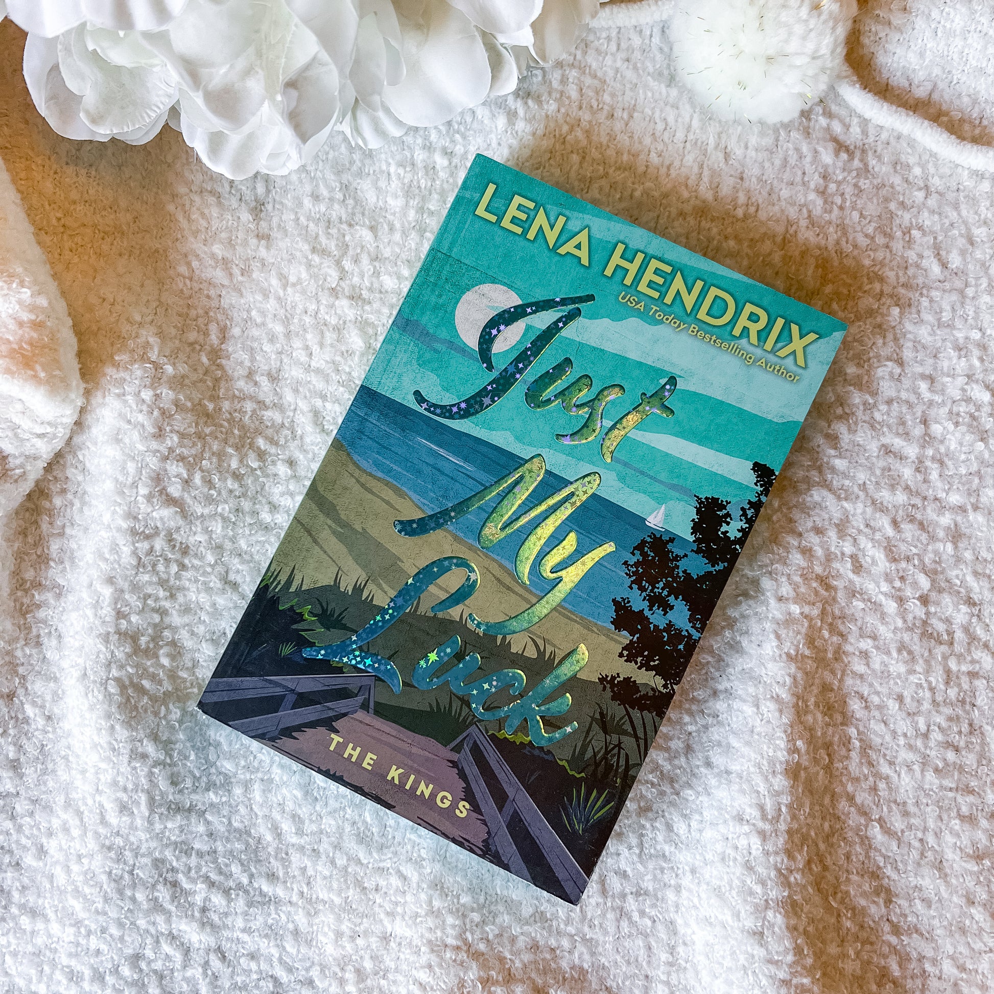 Book titled 'Just My Luck' by Lena Hendrix on a textured surface with flowers.