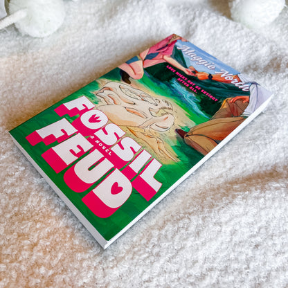 A paperback book titled 'Fossil Feud' by Maggie Nelson lies diagonally on a soft, textured white blanket. The cover features a vibrant illustration with a prominent green background, bold pink and white title text, and a cartoonish beige fossil character.
