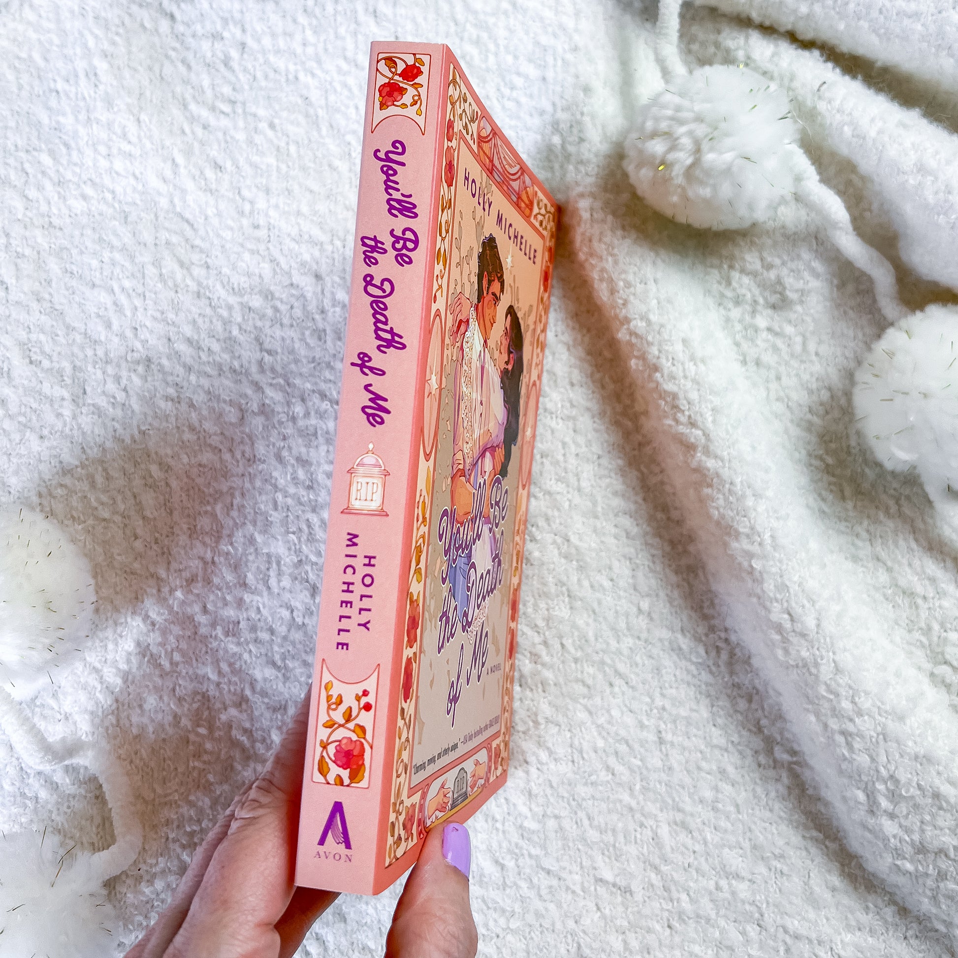 A hand holds a pink book titled 'You'll be the death of me' by Hollie Michelle. The book cover features a colorful illustration of a girl and features the Avon logo at the bottom.