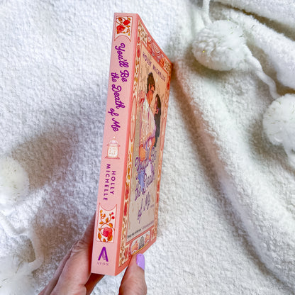 A hand holds a pink book titled 'You'll be the death of me' by Hollie Michelle. The book cover features a colorful illustration of a girl and features the Avon logo at the bottom.