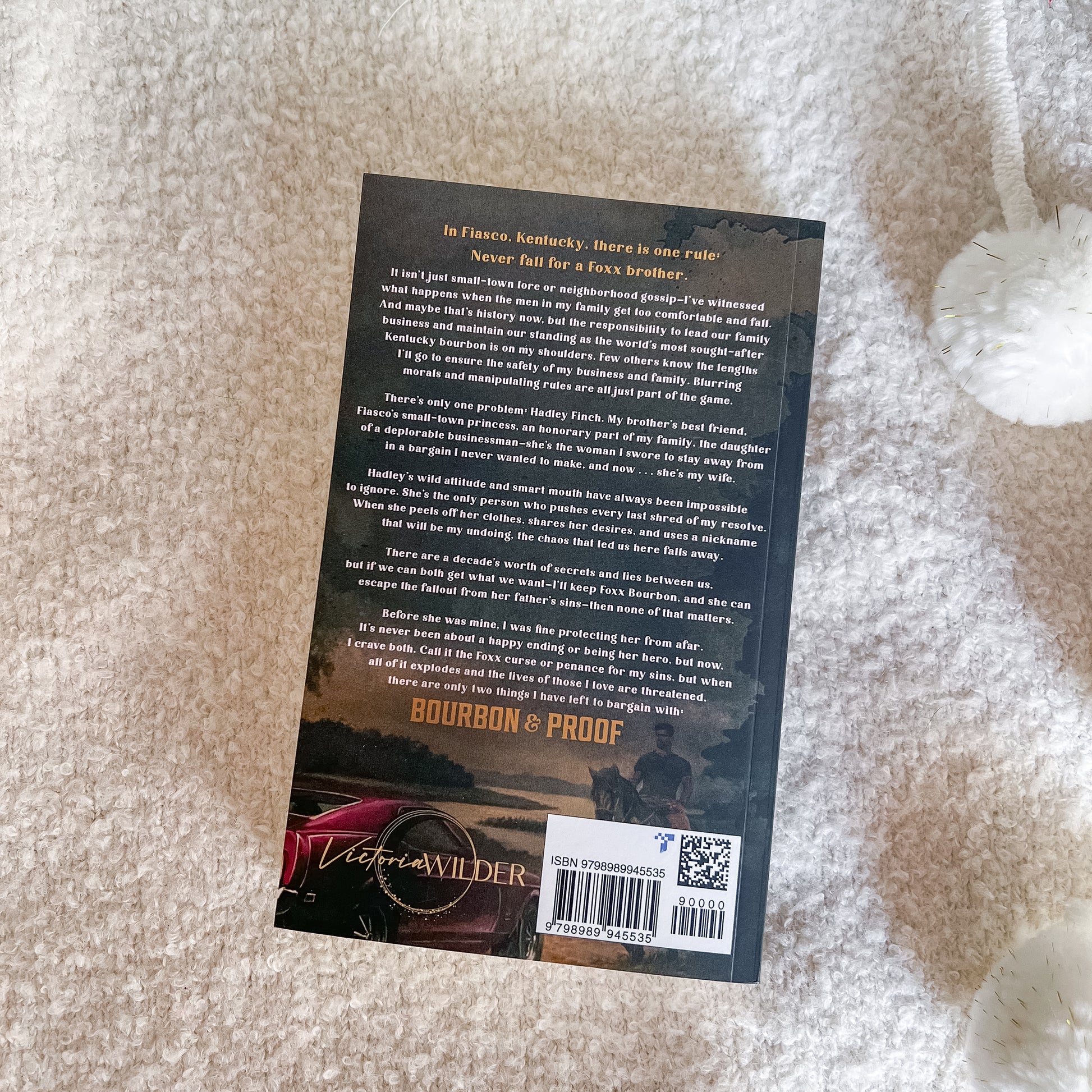 The back cover of the book 'Bourbon & Proof' by Victoria Wilder, photographed flat on a textured cream-colored blanket. The cover features a dark landscape with a red car and a silhouette of a man, with the title and author name clearly visible.