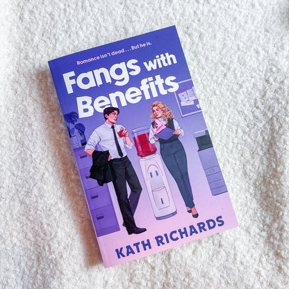 A paperback book titled 'Fangs with Benefits' by Kath Richards, featuring a purple and pink cover with an illustration of a man and woman in an office setting, photographed on a white textured fabric background.