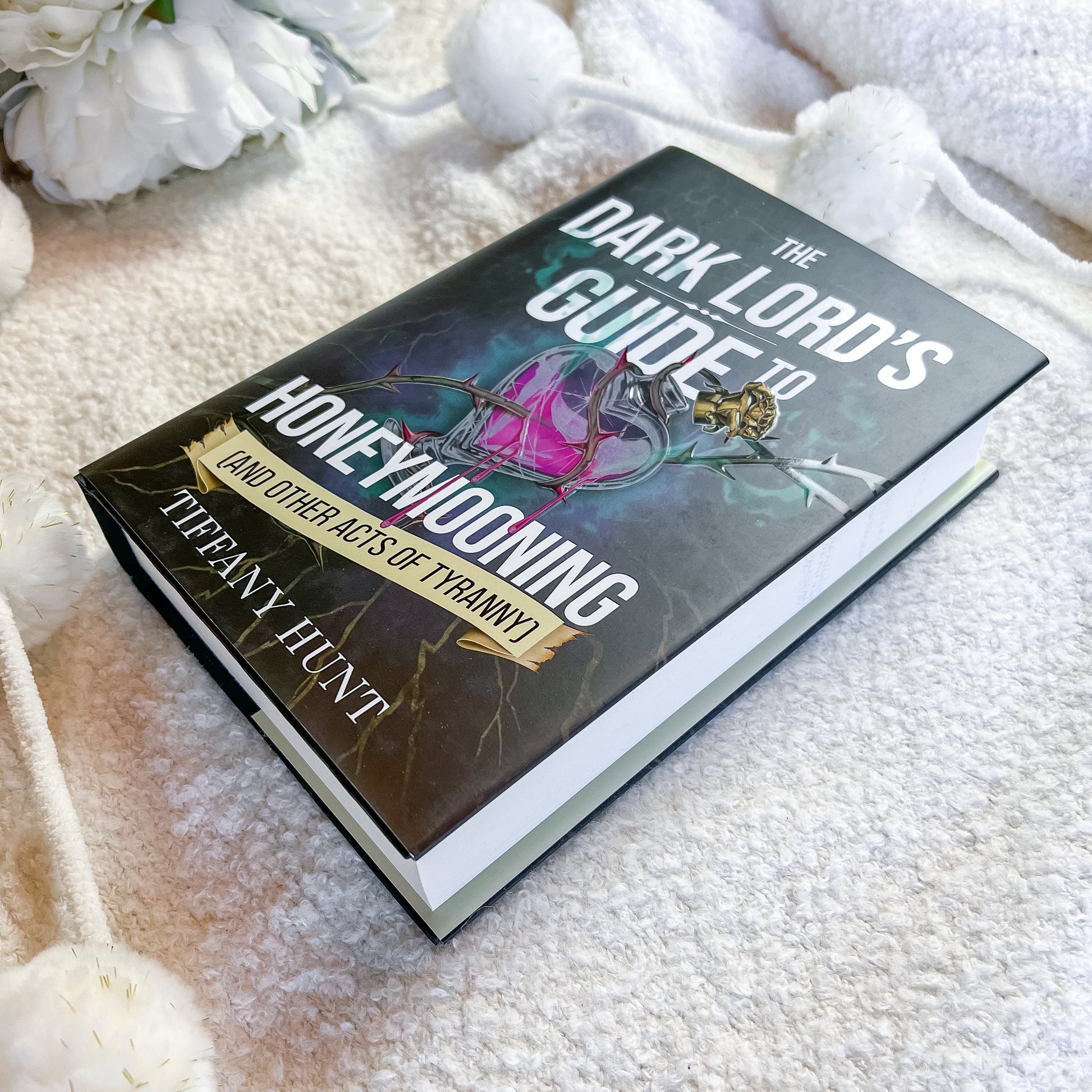 A hardcover book titled 'The Dark Lord's Guide to Honeywooning' by Tiffany Huntz, featuring a dark cover with a pink and purple glass bottle illustration, placed on a soft white textured blanket with white pom-pom tassels and white flowers.