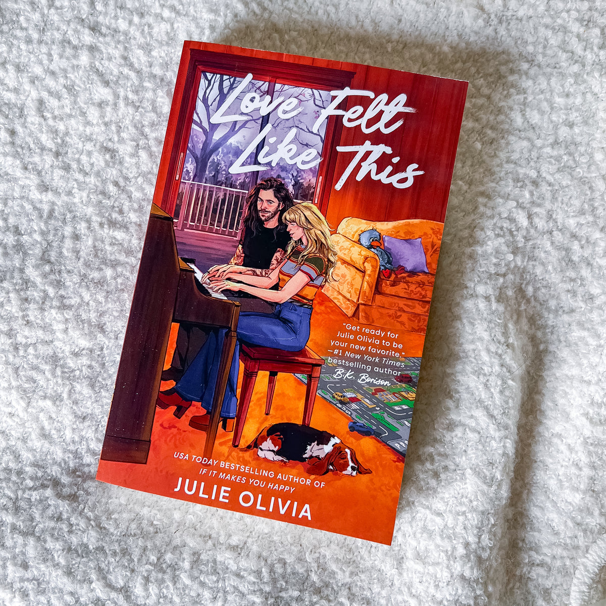 A paperback copy of the book 'Love Felt Like This' by Julie Olivia lies on a white textured blanket. The cover features a warm illustration of a woman playing piano with a man, set against a red and orange background with a dog sleeping on the floor.
