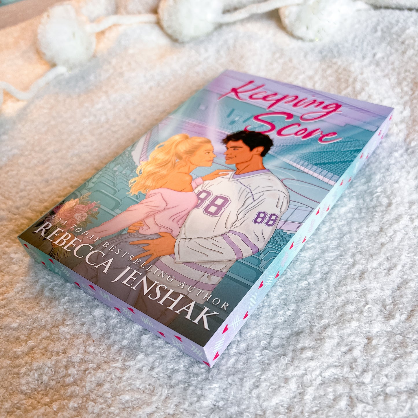 A paperback book titled "Keeping Score" by Rebecca Jenshak, featuring an illustrated cover of a blonde woman and a man in a white sports jersey, resting on a soft white textured blanket.