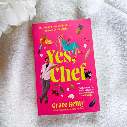 A hardcover book titled 'Yes, Chef' by Grace Reilly lies flat on a textured white fabric surface with white flowers in the background. The cover is bright pink with large yellow title text and colorful kitchen-themed illustrations.