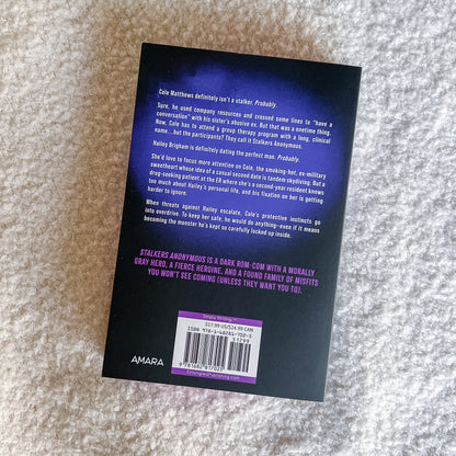 The back cover of a book titled 'Stalkerers Anonymous' by Coe Matthews, photographed flat on a textured gray fabric background. The cover is black with white and purple text and features a barcode with Amara branding at the bottom.
