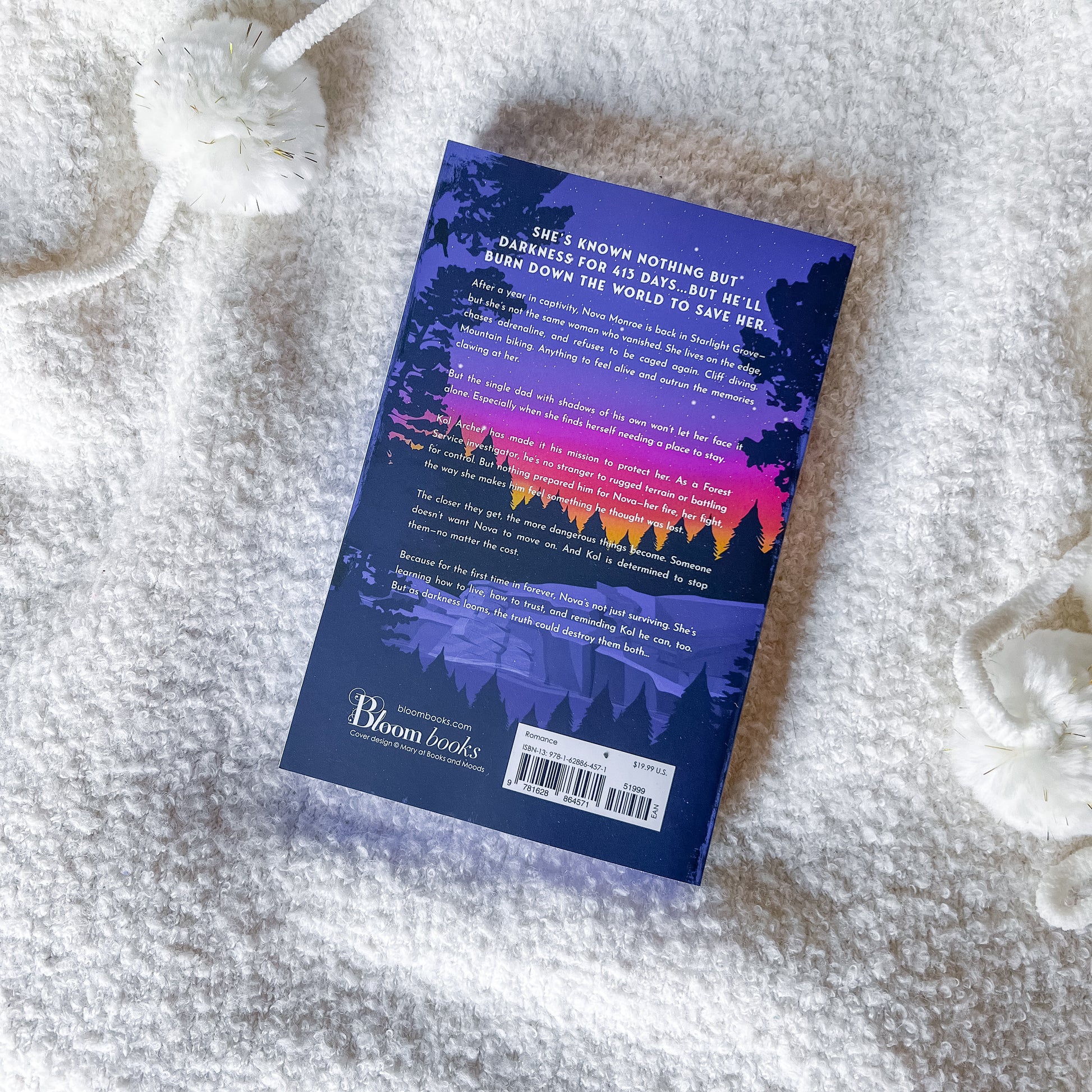 The back cover of a book by Bloom Books, featuring a purple and pink sunset forest design, laid flat on a white textured blanket with white knit accessories.