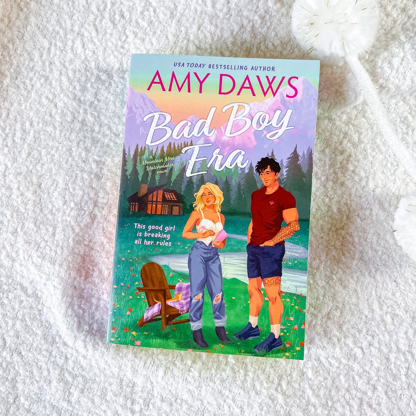 A book titled "Bad Boy Era" by Amy Daws lies on a white textured blanket. The cover features an illustrated scene with a blonde woman in ripped jeans and a man in a red shirt, set against a backdrop of mountains, a cabin, and a lake.