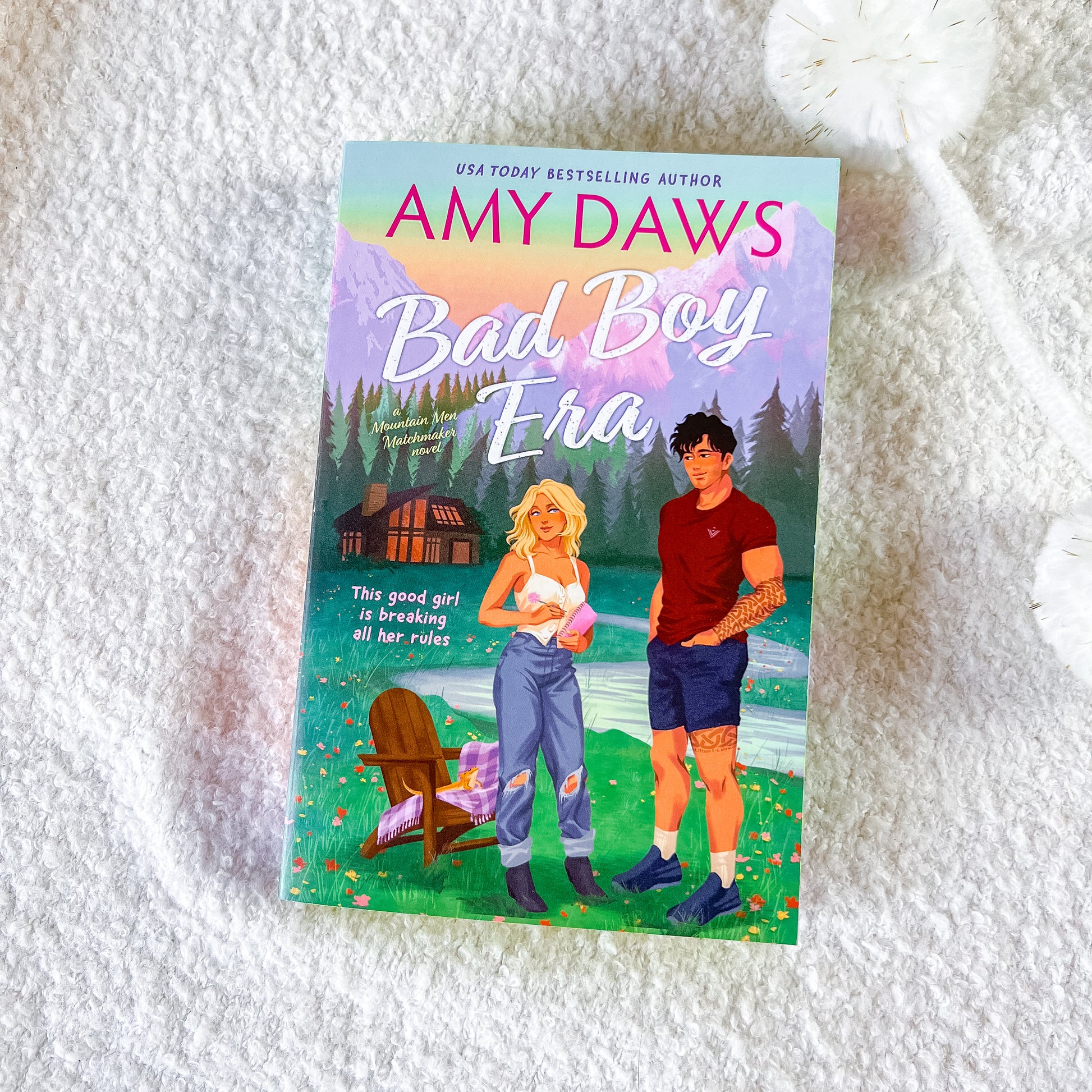 A book titled "Bad Boy Era" by Amy Daws lies on a white textured blanket. The cover features an illustrated scene with a blonde woman in ripped jeans and a man in a red shirt, set against a backdrop of mountains, a cabin, and a lake.