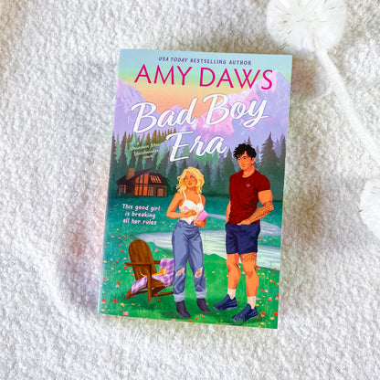 A book titled "Bad Boy Era" by Amy Daws lies on a white textured blanket. The cover features an illustrated scene with a blonde woman in ripped jeans and a man in a red shirt, set against a backdrop of mountains, a cabin, and a lake.
