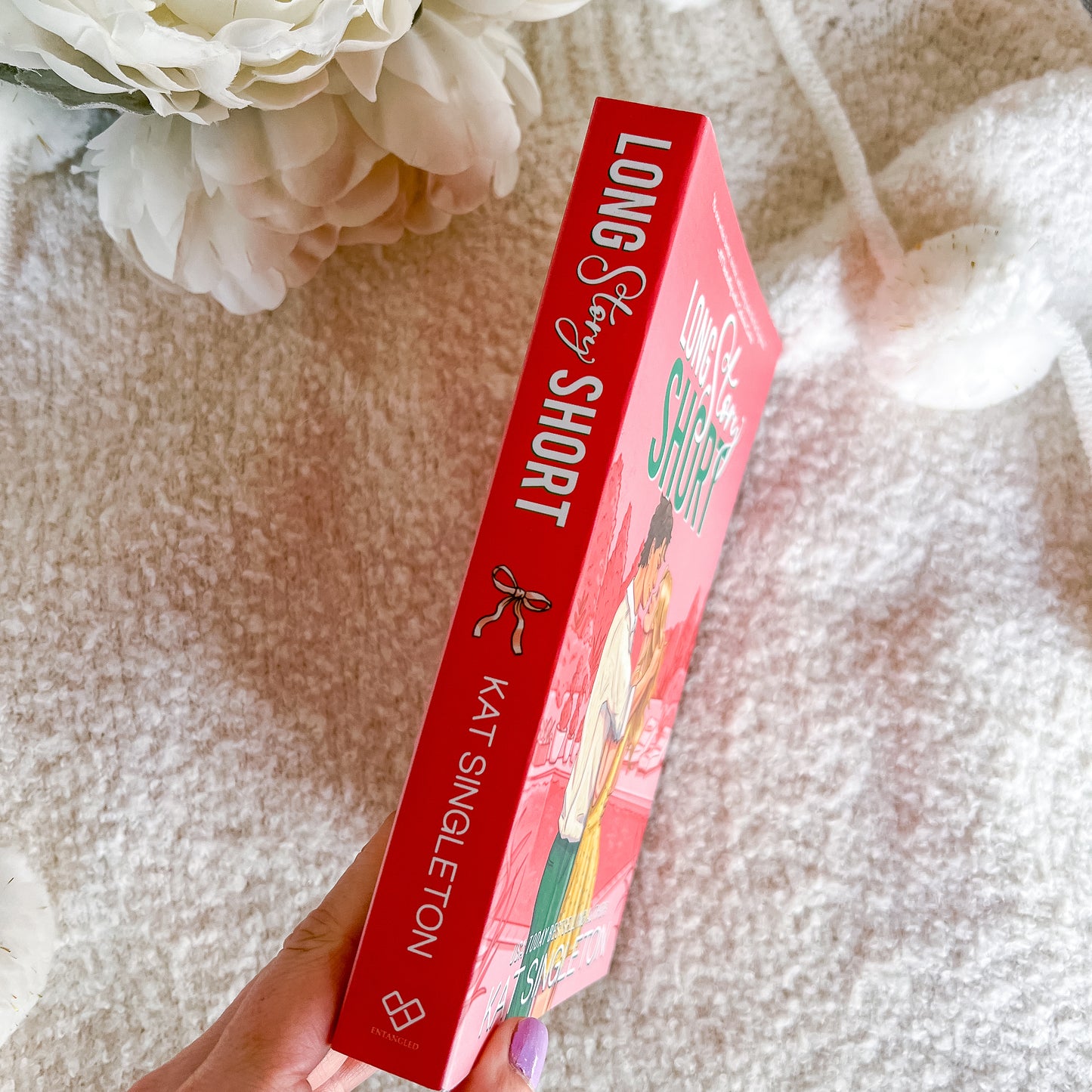 A hand holds a copy of the book 'Long Story Short' by Kat Singleton, showing the red spine and front cover, set against a soft beige textured blanket with white flowers in the background.