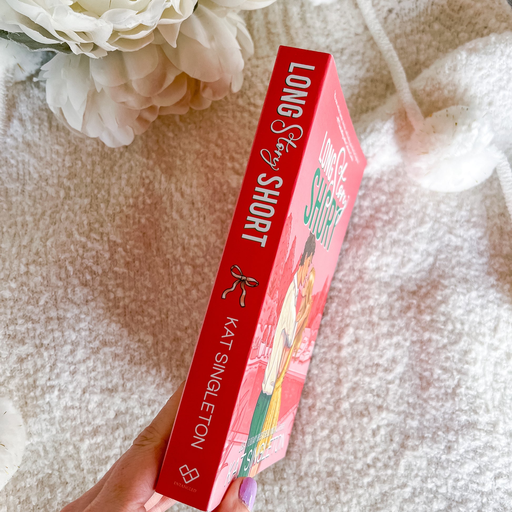 A hand holds a copy of the book 'Long Story Short' by Kat Singleton, showing the red spine and front cover, set against a soft beige textured blanket with white flowers in the background.