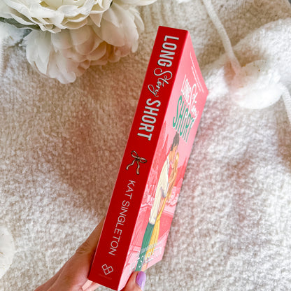 A hand holds a copy of the book 'Long Story Short' by Kat Singleton, showing the red spine and front cover, set against a soft beige textured blanket with white flowers in the background.