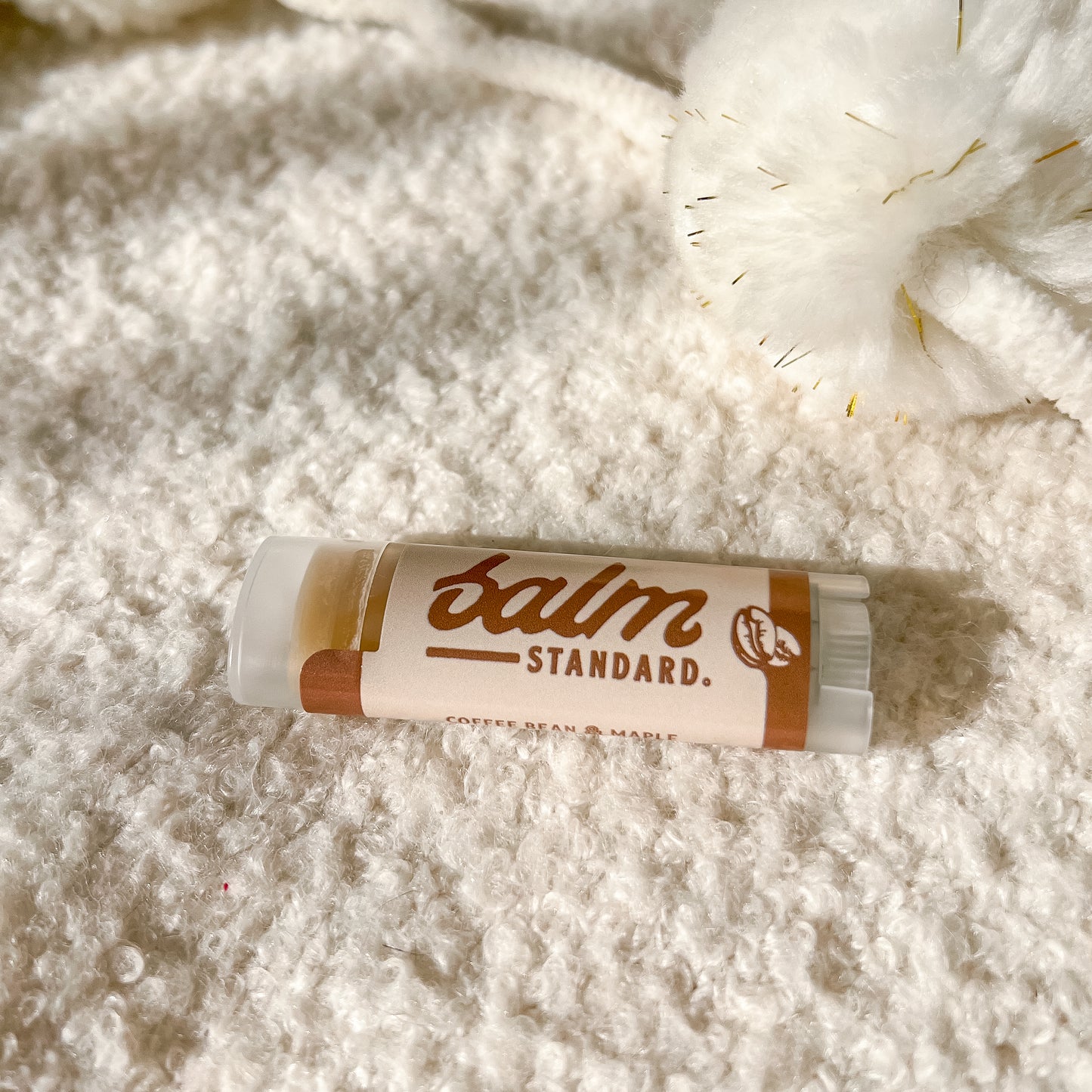 A tube of Balm Standard lip balm lies on a soft, textured white blanket with a fluffy white pom-pom nearby. The label is cream-colored with brown text and accents.